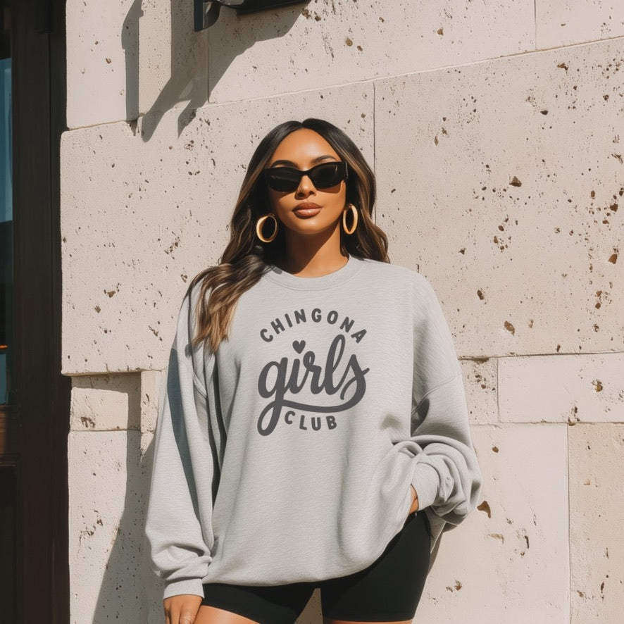 Woman wearing a gray sweatshirt with 'Chingona Girls Club' text, standing against a stone wall.