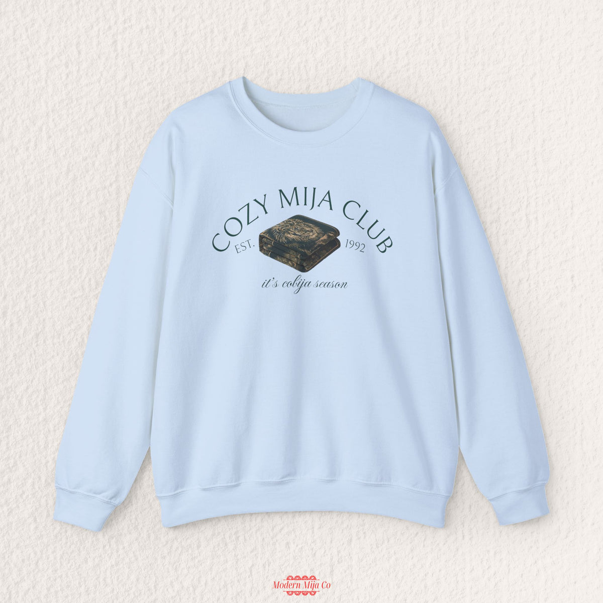 A light blue crewneck sweatshirt with a graphic on the chest reading 'COZY MIJA CLUB EST. 1992' and 'it's cozy season,' featuring a dark camouflage-patterned box illustration.